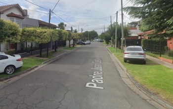 Vista de calle asfaltada con casas de dos pisos, árboles en las aceras, automóviles estacionados y tendido eléctrico aéreo
