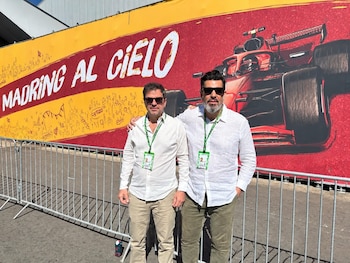 Dos hombres con camisas blancas y gafas de sol posan ante un panel con el texto Madring al cielo y el dibujo de un coche rojo de carreras