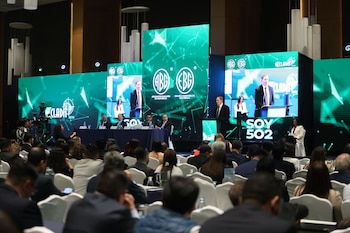 Vista frontal de una conferencia con un orador en el escenario, flanqueado por grandes pantallas que muestran logos bancarios y un público asistente