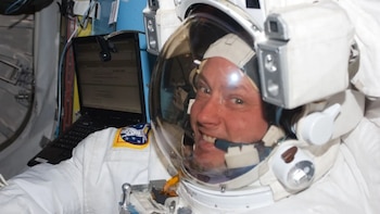Astronauta Michael Fincke con traje espacial blanco, casco transparente y una insignia. Una laptop y cableado se ven en el fondo