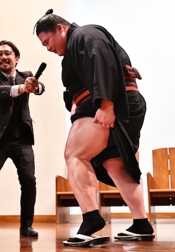 Ura Kazuki levanta el kimono y muestra la musculatura de su pierna izquierda; un presentador con traje sostiene un micrófono frente a él