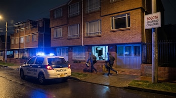 Patrulla policial con luces azules en calle mojada de noche. Dos encapuchados corren con bolsas frente a una vivienda de ladrillo con ventana rota. Cartel 'Bogotá - Seguridad Ciudadana'.