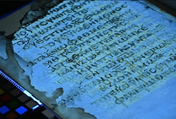 Manuscrito antiguo con texto en griego bajo luz UV, revelando letras amarillentas sobre fondo azul. La página muestra desgaste y un gráfico de calibración