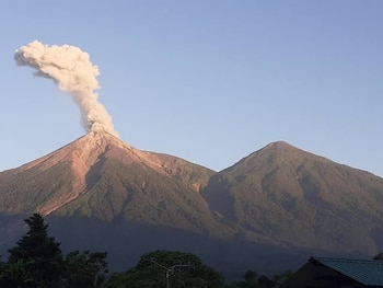 Un volcán que expulsa humo y ceniza junto a otra montaña, con árboles y el tejado de una estructura en la parte inferior