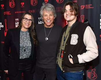 Tres personas posan sonrientes. Una mujer de cabello largo y gafas, un hombre de cabello gris y un joven con cabello largo y chaqueta tipo universitaria