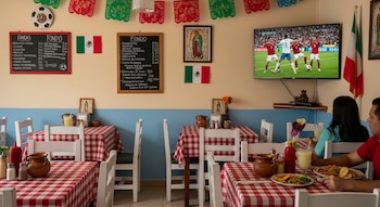 Fonda mexicana con manteles a cuadros, decoraciones patrias y TV transmitiendo fútbol. Clientes observan el partido mientras disfrutan de la comida típica.