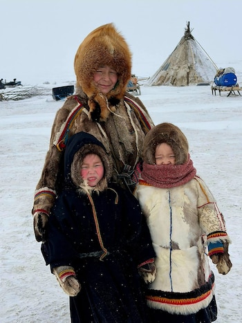 Una adulta y dos niños Nenets, vestidos con abrigos de piel y capuchas, posan sonrientes en un paisaje nevado. Al fondo, una tienda cónica y trineos