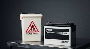 Batería Casely Power Pod E33A blanca y negra con terminales en una mesa metálica, junto a un contenedor crema con un símbolo de residuo peligroso (llama tachada).