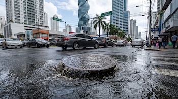 Una tapa de alcantarilla con la inscripción IDAAN Panamá rebosa agua en una calle mojada rodeada de vehículos y rascacielos.