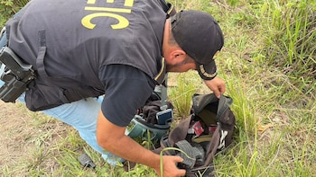 Agente con gorra y chaleco oscuro inspecciona un bolso abierto en el suelo. Se observan cargadores de arma, cajas de munición, un celular y un radio en la hierba
