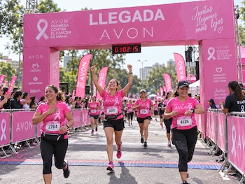 Varias mujeres con camisetas rosadas cruzan la meta debajo de un arco rosado con letreros sobre la concientización del cáncer de mama