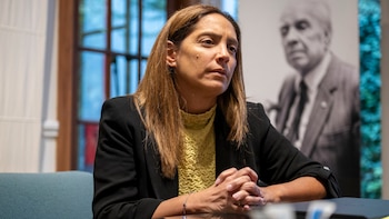 Mujer de cabello castaño con blazer negro y camisa amarilla texturizada, sentada con las manos entrelazadas; un retrato borroso de Borges se ve detrás