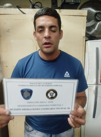 Un hombre de cabello oscuro y camiseta azul sostiene un documento oficial de la Policía de la Ciudad. Detrás se aprecian un mueble beige y un ventilador