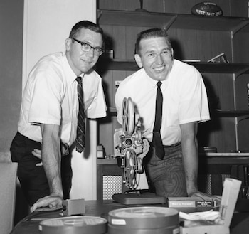 Una foto en blanco y negro de dos hombres, Raymond Berry y Johnny Unitas, sonriendo junto a un proyector de cine y rollos de película sobre una mesa