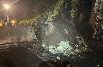 Imagen nocturna de una carretera bloqueada por una roca grande y escombros de un deslizamiento. Un cargador frontal se ve a la izquierda, limpiando la vía