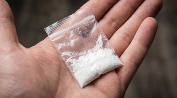 Primer plano de una mano sosteniendo una pequeña bolsa de plástico transparente cerrada con cierre hermético. Dentro de la bolsa hay una sustancia blanca cristalina o en polvo.