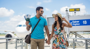 Pareja latina sonriente de la mano en una pasarela de embarque, con pasaportes y boletos, frente a un avión blanco bajo un cielo azul.