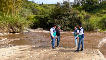 Cuatro hombres con chalecos de seguridad y ropa informal inspeccionan una vía de tierra junto a un río de aguas marrones. Al fondo, colinas verdes y vegetación