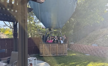 Un globo aerostático azul claro con una canasta de mimbre está aterrizado en un patio trasero. Dentro de la canasta, varias personas sonríen y saludan