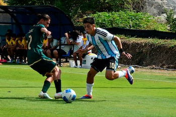 Dos jugadores de fútbol, uno con camiseta celeste y blanca de Argentina y otro con camiseta verde de Bolivia, disputan un balón blanco y azul en un campo verde