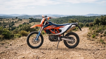 Motocicleta de enduro naranja, azul y blanca estacionada sobre un camino de tierra y piedras entre colinas y vegetación.