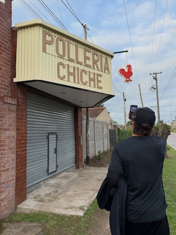 Un hombre de espaldas con camiseta y gorra negras toma una foto con su teléfono a la marquesina de un local llamado Pollería Chiche