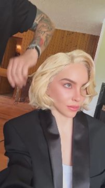 Mujer de cabello rubio, corto y ondulado, recibe un peinado de un estilista con brazos tatuados. Viste chaqueta negra y top blanco