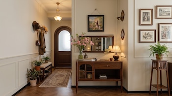 Pasillo interior con puerta de madera arqueada, perchero con sombreros y chaqueta, banco, macetas, alfombra, cómoda con espejo, lámpara, flores y cuadros.