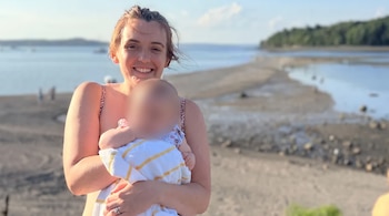 Una mujer sonriente sostiene en sus brazos a un bebé con el rostro difuminado en una playa con colinas y vegetación de fondo.