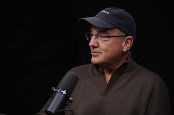 Primer plano de un hombre con gorra azul oscura con el logo ALPHA, gafas y un suéter marrón oscuro. Un micrófono negro en primer plano inferior izquierdo. Fondo negro