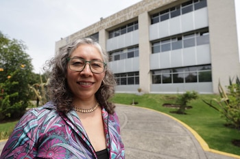 Una mujer con gafas y camisa de tonos rosados sonríe frente a un edificio institucional con jardín