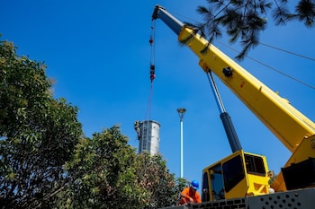 Una grúa amarilla sostiene una estructura metálica cilíndrica rodeada de árboles y bajo un cielo despejado