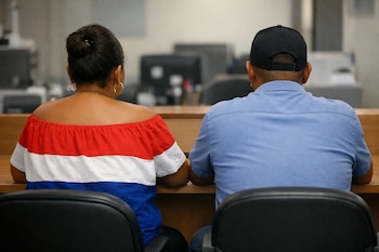 Vista trasera de dos adultos sentados en un escritorio. La mujer lleva una blusa con franjas rojas, blancas y azules, y el hombre una gorra negra.