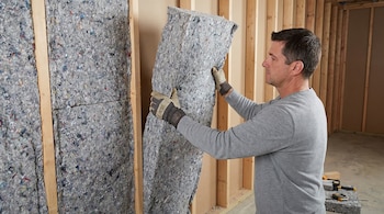Un hombre con guantes instala una manta de aislamiento gris con motas de colores en una pared con estructura de madera. Hay herramientas en el suelo.