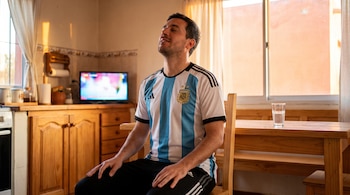 Hombre con camiseta de la selección argentina con tres estrellas, sentado en una silla de madera en una cocina con una televisión y un vaso de agua.