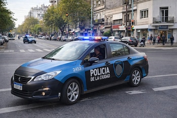 Un automóvil patrullero de la Policía de la Ciudad de Buenos Aires con balizas encendidas en una calle urbana con edificios, árboles y personas.