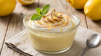 Un cuenco de vidrio con mousse de limón amarillo pálido, cubierto con merengue italiano tostado, ralladura de limón y hojas de menta fresca, sobre un paño y madera.