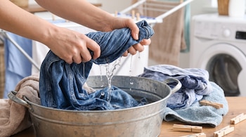 Manos exprimiendo prenda de mezclilla azul sobre un barreño metálico. Agua gotea. Ropa mojada, toallas y pinzas de madera en la mesa. Lavadora al fondo.