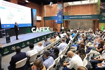 Mujer exponiendo en un escenario frente a una pantalla con mapas y texto, con una audiencia sentada en sillas blancas. Logotipos de CAFEX y ANACAFÉ