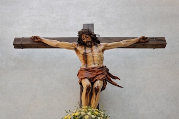 Estatua de Cristo crucificado en cruz de madera con corona de espinas, sangre, paño de pureza y base con flores blancas y amarillas. Fondo blanco.