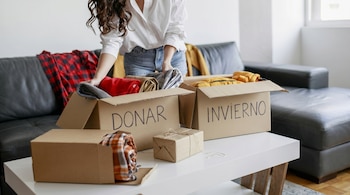 Una persona con camisa blanca dobla y coloca ropa en dos cajas de cartón sobre una mesa. Las cajas están etiquetadas con 'DONAR' e 'INVIERNO' en mayúsculas negras.