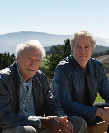 Dos hombres, Clint Eastwood de cabello blanco y Kyle Eastwood de cabello rubio, posan sentados al aire libre con un paisaje de montañas y árboles