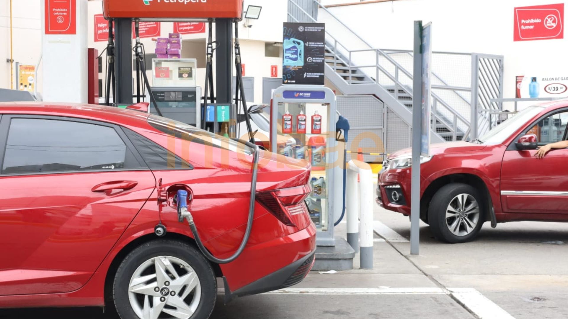 Un coche sedán rojo es abastecido de combustible en una estación de servicio Petroperú, con una manguera azul conectada al tanque mientras un SUV rojo espera en segundo plano. (Paula Elizalde / Infobae Perú)