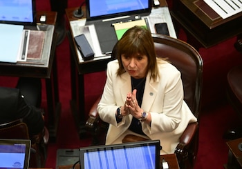 Vista superior de legisladores en una cámara del senado. Un hombre con barba y una mujer rubia están de pie. Monitores y documentos sobre mesas de madera oscura