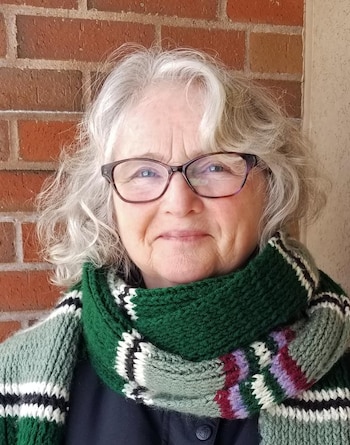 Una mujer de cabello canoso y gafas sonríe mientras viste una bufanda tejida de color verde ante un muro de ladrillo