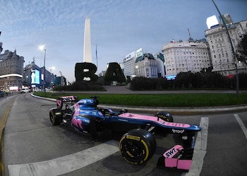 Coche de carreras azul y rosa de Franco Colapinto estacionado en una calle de Buenos Aires, con el Obelisco, la escultura "BA" y edificios urbanos de fondo bajo un cielo nublado