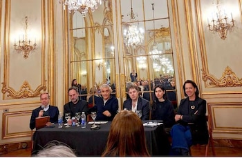 Seis personas sentadas ante una mesa con manteles oscuros y copas, en un salón con espejos altos, arañas y molduras doradas