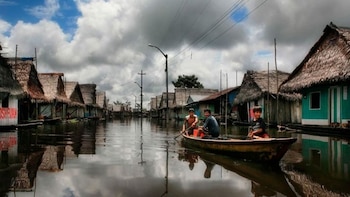 Tres personas viajan en una canoa de madera por una calle de agua flanqueada por casas sobre pilotes y postes de electricidad