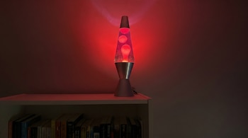 Una lámpara de lava roja encendida, con burbujas ascendiendo, está colocada sobre un estante blanco con libros y una pared roja brillante.
