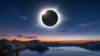 Eclipse solar total con un halo brillante en el cielo azul oscuro. Debajo, un lago tranquilo con una pequeña isla y montañas escarpadas bajo un horizonte de tonos naranjas y morados.